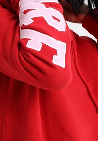 Felpa rossa con una texture morbida, caratterizzata da un grande numero bianco stampato sulla manica. Il design ha un'estetica sportiva e casual.