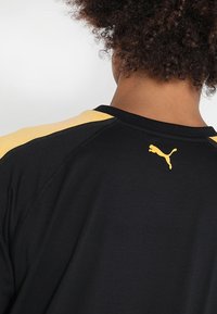 Svart sporttröja med gula axel detaljer och en gul Puma-logga vid nacke. Tillverkad av mjukt, andningsbart material.