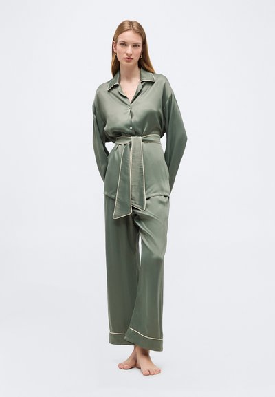 Γυναίκα στέκεται ξυπόλυτη, φορώντας χαλαρή, σατέν πιτζάμα σε ελιά πράσινο χρώμα με λευκό περιθώριο και δεμένη ζώνη στη μέση.