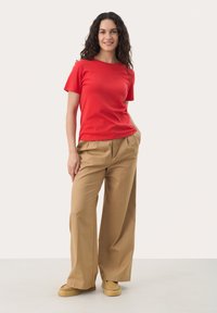 T-shirt rouge à manches courtes associé à un pantalon beige ample et des chaussures slip-on marron clair, sur un fond neutre.