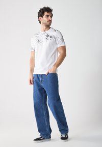 White t-shirt with black floral print on shoulders, paired with loose blue denim jeans and black sneakers, standing against a light background.