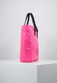 Růžová taška přes rameno s černými uchy, vyrobená z texturované látky. Na jedné straně má potištěné logo a nabízí prostorný, vzpřímený design.
