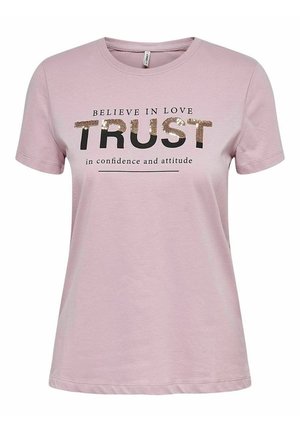 Persoon draagt een roze T-shirt met de tekst "TRUST" en versleten skinny blauwe jeans, staat met handen in de zakken en draagt nude sandalen met hakken.