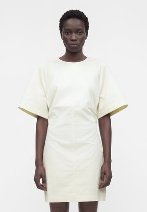 Femme aux cheveux courts portant une robe crème à manches courtes, longueur genou, debout devant un fond blanc uni.