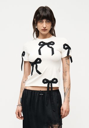 Donna che indossa una camicia bianca aderente con più fiocchi neri e una gonna nera con dettaglio in pizzo, con un braccialetto al polso sinistro.