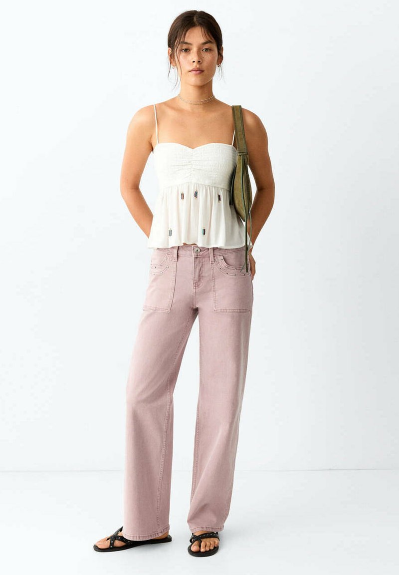 Femme debout portant un haut blanc sans manches, un pantalon large rose clair, des sandales noires et un sac à bandoulière vert olive.
