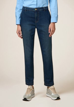 Kobieta ubrana w granatowe jeansy o prostych nogawkach, jasnoniebieską koszulę z długim rękawem oraz beżowo-szare trampki, stojąca na neutralnym tle.