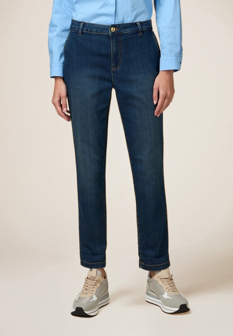 Femme portant un jean droit bleu foncé, une chemise à manches longues bleu clair et des baskets beige et grises, debout devant un fond neutre.