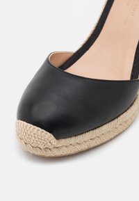 Chaussure espadrille en cuir noir avec bout arrondi, semelle enveloppée de jute et bride arrière élastique sur fond blanc.
