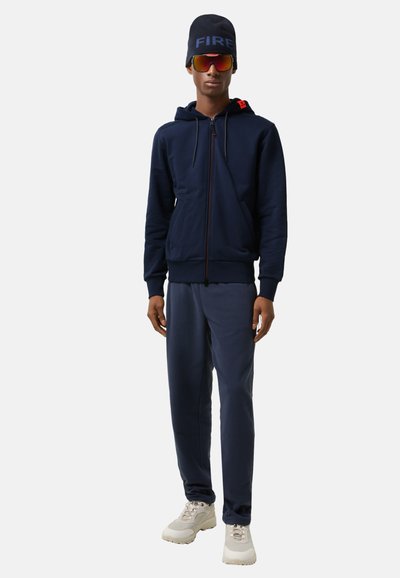 Navy zip-up hoodie with red accents, paired with matching sweatpants. Footwear is light-colored with a chunky design; accessories include a black beanie and reflective sunglasses.