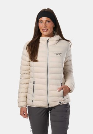 Femme portant une veste matelassée beige clair avec fermeture éclair frontale et poches, pantalon gris foncé et un bandeau noir, debout devant un fond blanc.