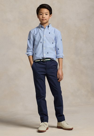 Chłopiec stoi z rękami w kieszeniach, ubrany w jasnoniebieską koszulę na guziki z logo, granatowe spodnie, paskowany pasek i beżowe buty.