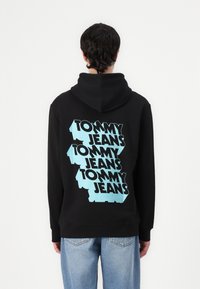 Zwarte hoodie met een groot blauw grafisch ontwerp waarop "TOMMY JEANS" in een herhalend patroon over de achterkant is afgebeeld. Voorzien van een trekkoord capuchon en geribde boorden.