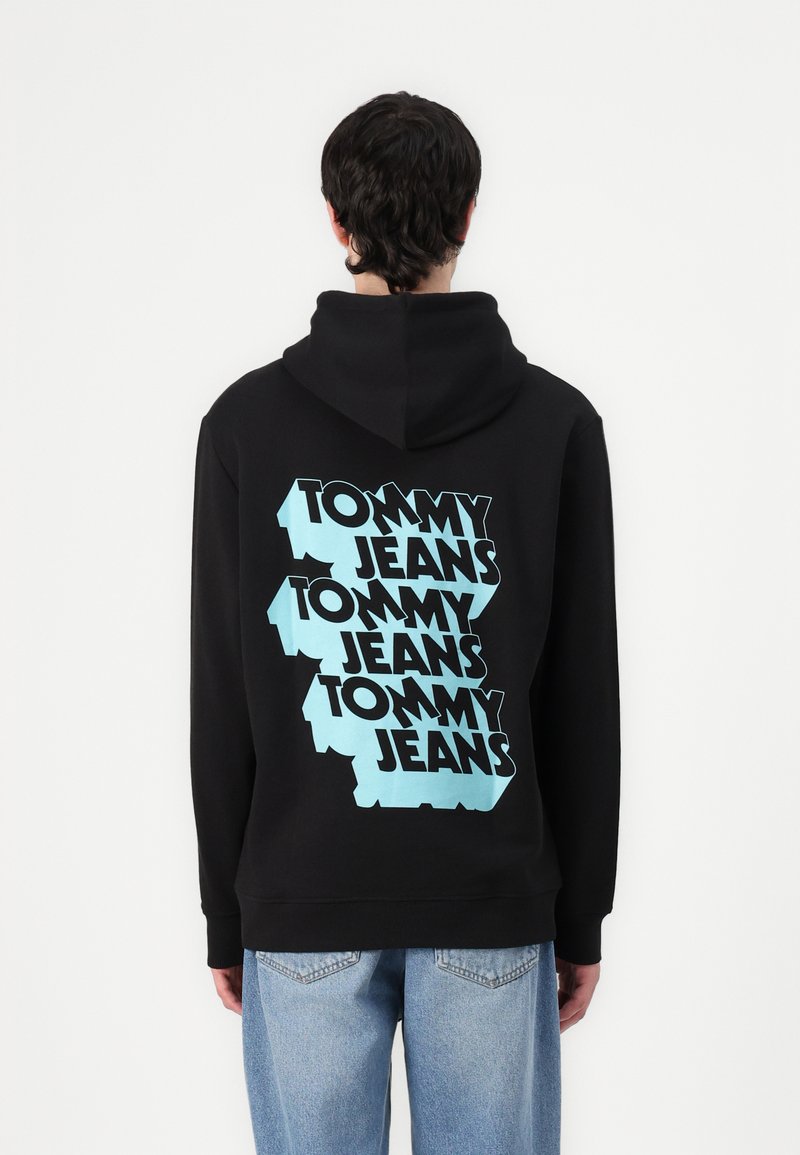 Zwarte hoodie met een groot blauw grafisch ontwerp waarop "TOMMY JEANS" in een herhalend patroon over de achterkant is afgebeeld. Voorzien van een trekkoord capuchon en geribde boorden.