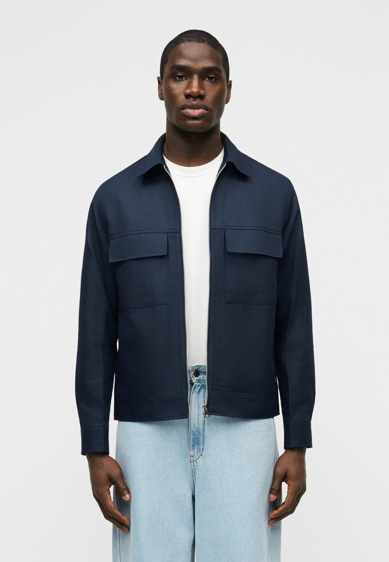 Νεαρός άνδρας φοράει μπλε μπουφάν με φερμουάρ πάνω από λευκό πουκάμισο και ανοιχτόχρωμα τζιν, στέκεται μπροστά σε απλό γκρι φόντο.