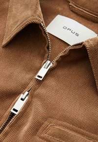 Brown corduroy jacket with two silver metal zippers partially zipped and a white clothing label reading "OPUS" inside the collar.