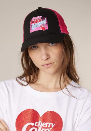 Jeune femme portant une casquette trucker Cherry Coke noire et rose ainsi qu'un t-shirt blanc avec un cœur rouge pailleté et le logo Cherry Coke.