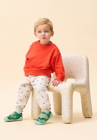 Felpa rossa, pantaloni beige a pois e scarpe verdi. Il bambino è seduto su una sedia chiara con una texture e bordi arrotondati. Design semplice e giocoso.