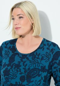 Robe teal avec un col rond et un motif floral paisley dans des teintes plus foncées. Accessoire féminin : grandes boucles d'oreilles créoles.