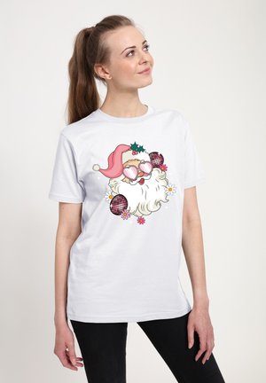 Kvinde iført en hvid T-shirt med et tegneserie-julemandsansigt i pink hjerteformede solbriller, omgivet af blomster og discokugler, kigger op og til højre.
