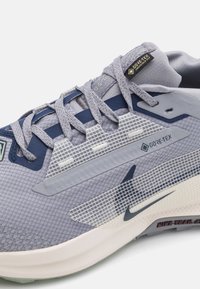 Scarpa da corsa GORE-TEX in grigio chiaro e blu navy. Presenta un tessuto testurizzato, dettagli riflettenti e una suola ammortizzata con un design a motivi.