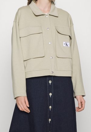 Beige jas met knopen, grote voorzakken en een logopatch, gedragen over een donkere rok met knopen.
