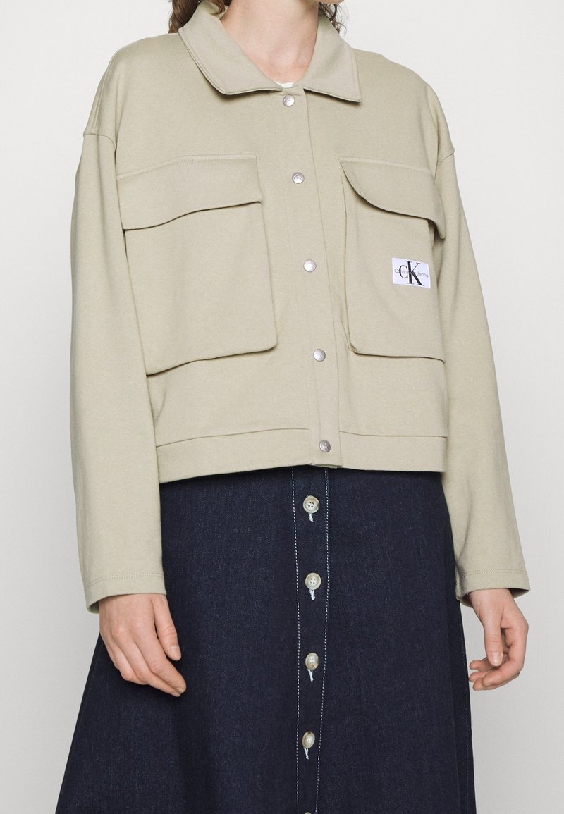 Veste beige boutonnée avec grandes poches frontales et écusson logo portée sur une jupe foncée boutonnée.