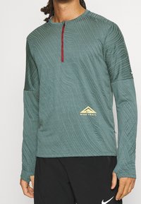 Long-sleeve teal running shirt with textured pattern, half-zip neckline, red zipper trim, and gold "Nike Trail" logo on the left side.