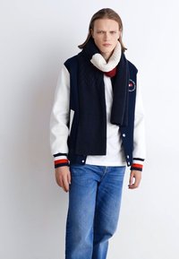 Navy and white letterman jacket with ribbed cuffs, worn over a white shirt; paired with a dark knitted scarf. Blue denim jeans complete the look.