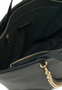 Borsa a mano nera con manico a catena dorata, zip superiore aperta che mostra una tasca interna con zip e fodera in tessuto.