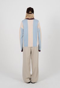 Personne vue de dos portant un pull rayé bleu pastel et beige, un pantalon beige à jambes larges, et une casquette de baseball noire.