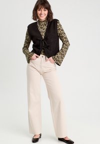 Gilet nero senza maniche con tessuto testurizzato, annodato davanti, sovrapposto a una maglietta a maniche lunghe verde con motivo leopardato. Pantaloni larghi beige chiaro.