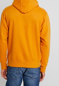 Hombre vestido con sudadera con capucha naranja y jeans azules, visto desde atrás contra un fondo claro y liso.