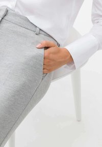 Osoba w jasnoszarych spodniach i białej koszuli z długim rękawem, z prawą ręką swobodnie umieszczoną w kieszeni, siedzi na białym krześle.