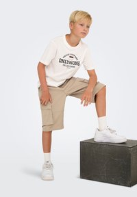 Shorts cargo beiges avec des poches latérales, associés à un T-shirt blanc avec un texte graphique noir. Baskets blanches et chaussettes côtelées à mi-mollet.