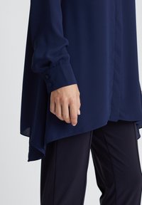 Marineblaue Bluse mit fließendem Design, langen Ärmeln und knopfartigen Manschetten. Der Stoff wirkt glatt und leicht. Kombiniert mit dunklen Hosen.