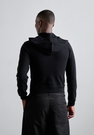 Black cropped hoodie made of smooth fabric, featuring a lined hood, ribbed cuffs, and a subtly branded hem. Worn with black pants.