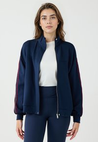 Giacca zip-up blu navy con colletto alto, tasche laterali e accenti a righe bordeaux sulle maniche. Realizzata in tessuto liscio ed elastico.