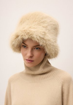 Žena v béžovém žebrovaném roláku a velké nadýchané béžové kožešinové čepici, která se mírně dívá stranou s neutrálním výrazem.