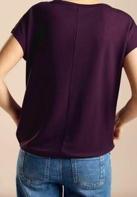 Blusa de manga corta en tejido morado oscuro, con un escote redondeado y un detalle de costura central en la parte posterior, combinada con jeans de color azul claro.