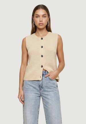 Kobieta w beżowej, bezrękawnikowej, dzianinowej kamizelce na guziki i jasnoniebieskich, wysokich dżinsach, stojąca z jedną ręką w kieszeni.