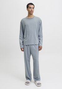 Man wearing loose-fitting gray long-sleeve top and matching pants, standing on a white background, looking slightly right.