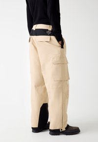 Beige Cargo-Hosen mit mehreren Taschen, einem schwarzen Bund und kontrastierenden schwarzen Akzenten am Saum. Hergestellt aus strapazierfähigem Stoff mit einer lässigen Passform.