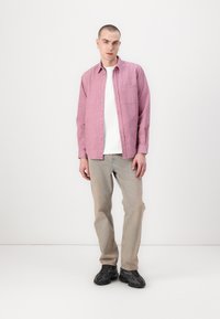 Pink button-up shirt with a pocket, worn over a white t-shirt. Light grey jeans and black sneakers complete the outfit. Minimalist background.