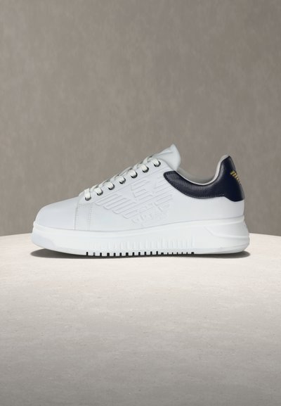 Λευκό sneaker χαμηλού προφίλ με μαύρη πίσω λωρίδα και ανάγλυφο λογότυπο στη πλευρά, τοποθετημένο σε φωτεινή επιφάνεια με φόντο απλό μπεζ.