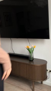 Holzfernsehschrank mit einer glatten, abgerundeten Oberseite und vertikalen Rillen, darauf eine durchsichtige Vase mit gelben und pinken Tulpen unter einem großen schwarzen Bildschirm.