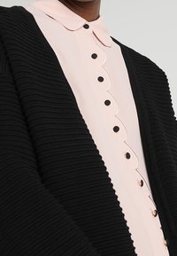 Svart ribbade cardigan över en ljusrosa blus med en vågig krage och svarta knappar fram.