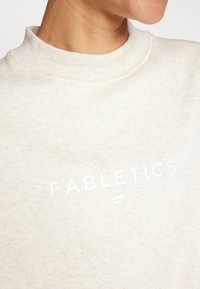 Svijetlosmeđi dukserica s visokim rebrastim ovratnikom, na prednjoj strani ispisana riječ "FABLETICS" bijelom bojom. Mekana, teksturirana tkanina.