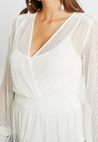 Top avvolgente in pizzo bianco con maniche lunghe, caratterizzato da una profonda scollatura a V e dettagli testurizzati. Vita smock per esaltare la vestibilità e il movimento.