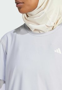 Camiseta atlética de color gris claro con tejido texturizado y escote redondeado, combinada con un hiyab drapeado de color crema. Detalle del logo en blanco visible.
